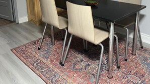 Un comedor con una mesa negra, cuatro sillas, un tapete estampado, un armario de madera con estantes y un reloj en la pared.