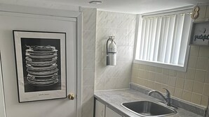 Una cocina pequeña con un lavamanos, un cuadro enmarcado en la puerta, un extintor de incendios en la pared y baldosas de suelo con diseño.