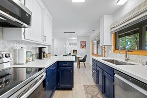 Une cuisine moderne avec des meubles blancs, un mur d’accent bleu, des appareils électroménagers en acier inoxydable et un coin repas offrant une vue sur l’extérieur.