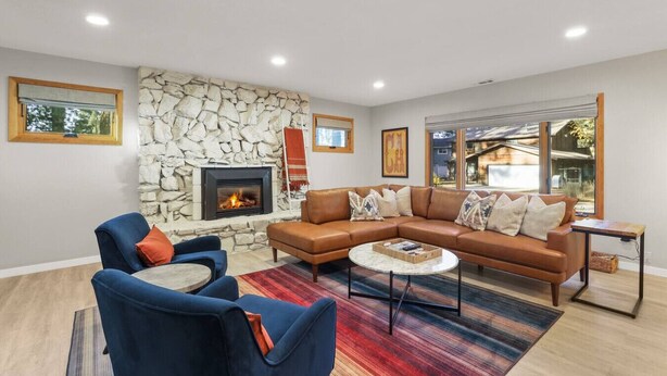 Una sala de estar con chimenea de piedra, un sofá de cuero marrón, sillones azules y un tapete colorido.