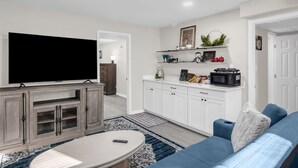 Een moderne woonkamer met een flatscreentelevisie, een blauwe bank, een salontafel, en een keukenhoek met witte kasten en een magnetron.