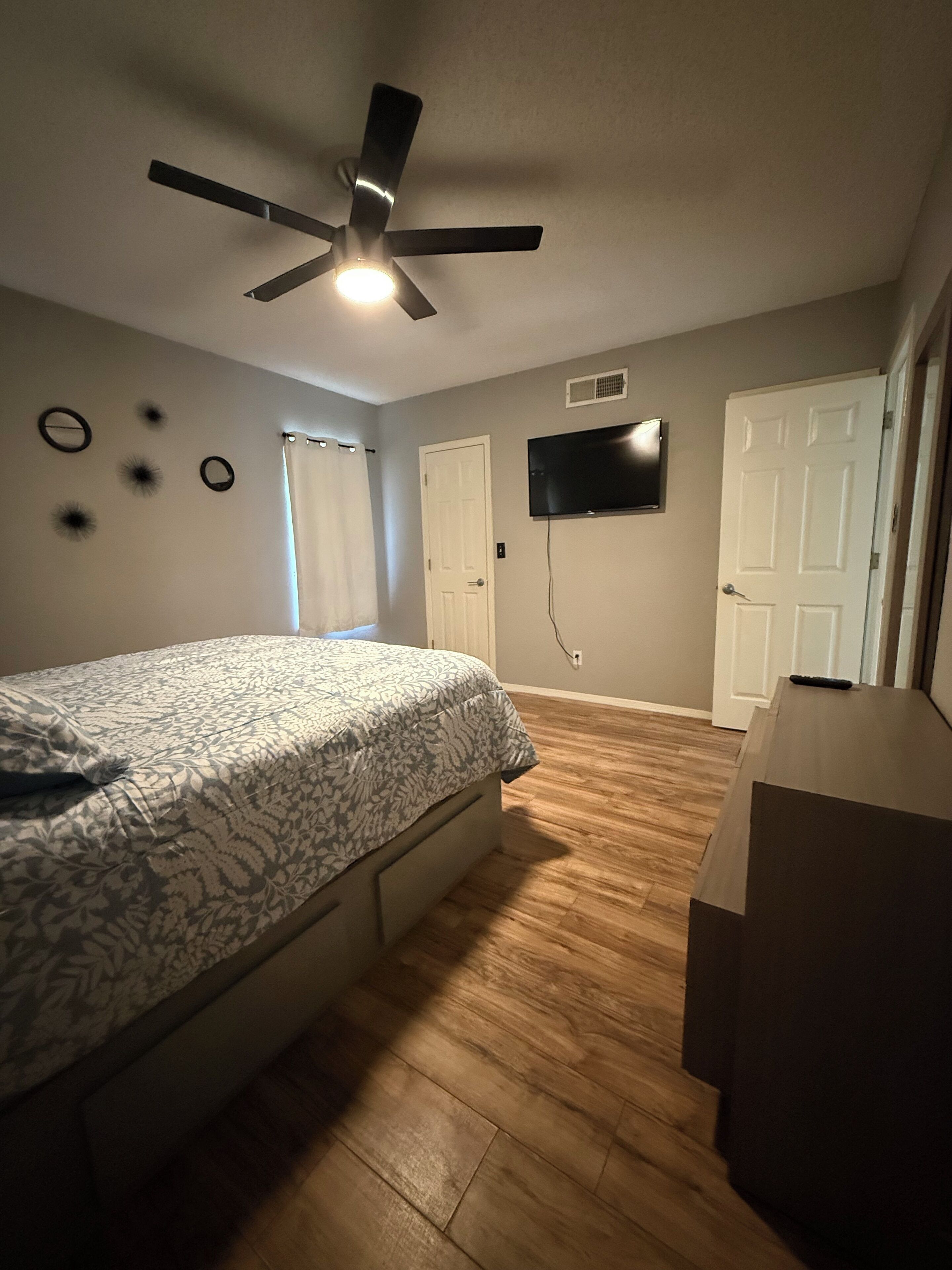 Quarto com cama, ventilador de teto, TV e piso de madeira.