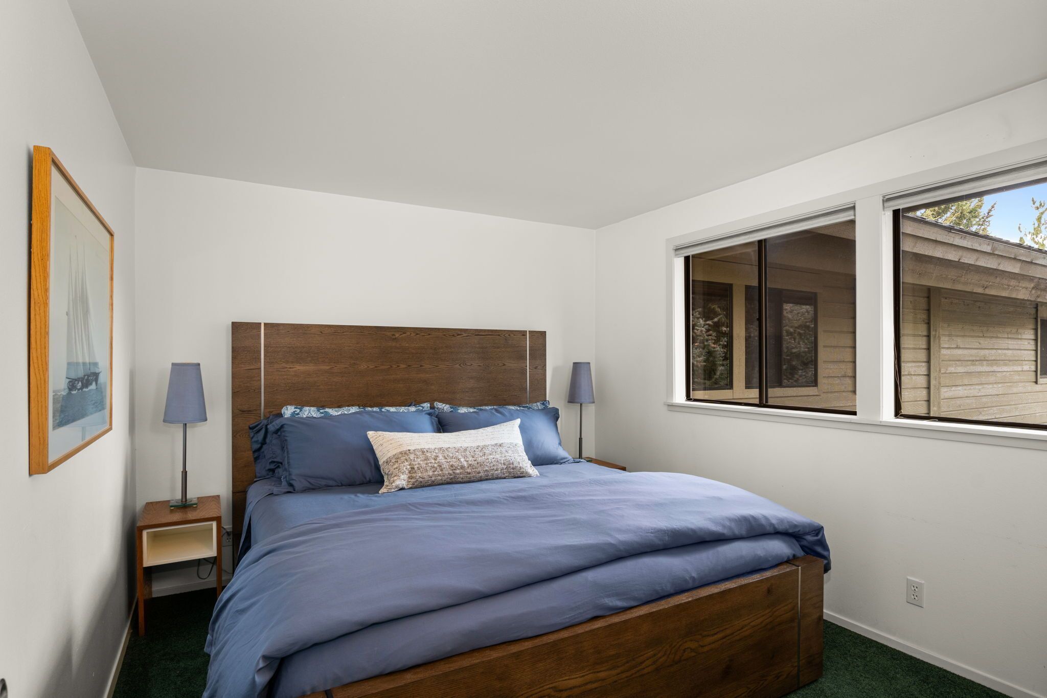Een slaapkamer met een houten hoofdbord, een enkel bed met blauw beddengoed, twee nachtlampjes, een ingelijst schilderij aan de muur en een raam met uitzicht op een gebouw.