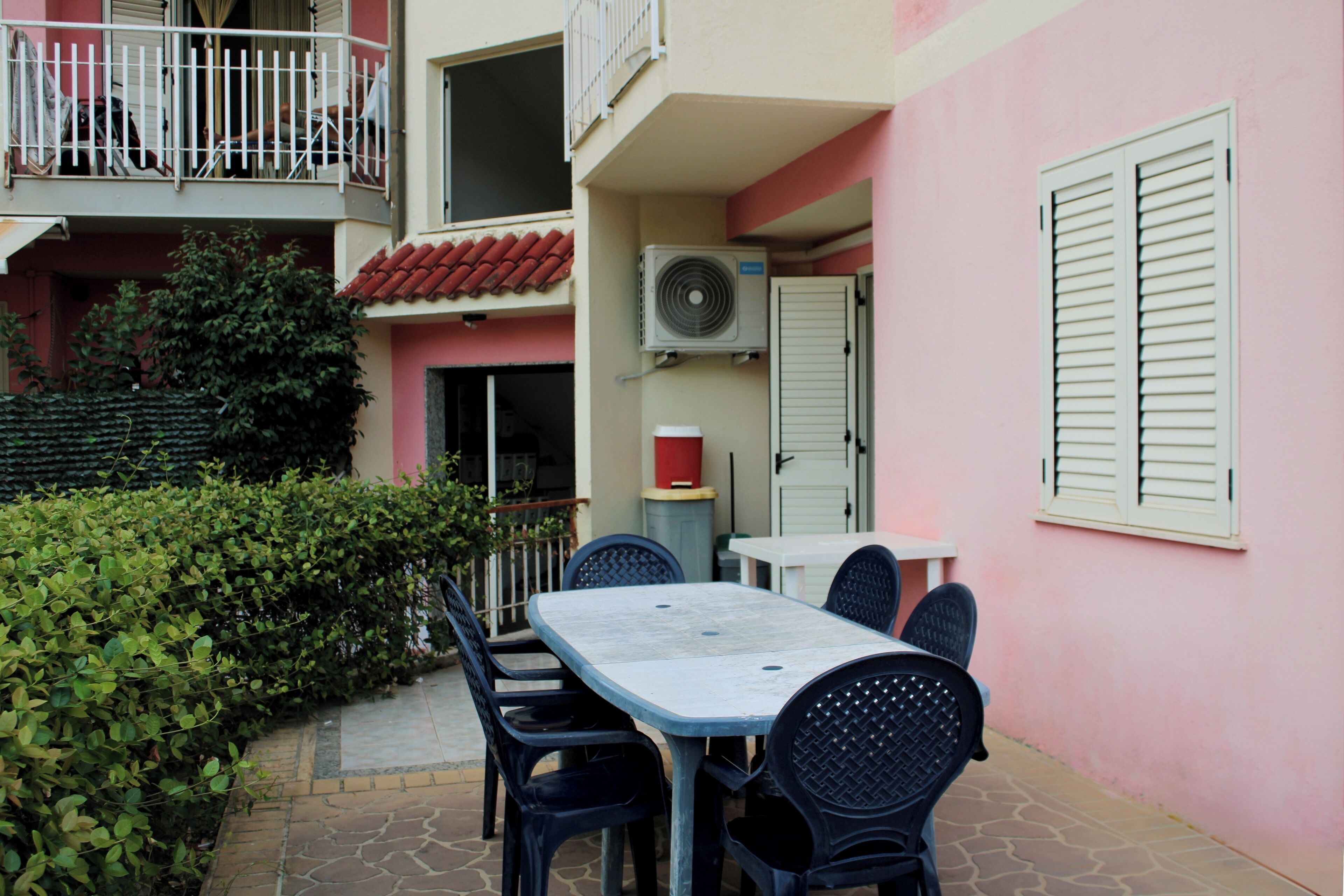 A patio with a table and chairs, a red-tiled roof, and a pink wall with shutters.