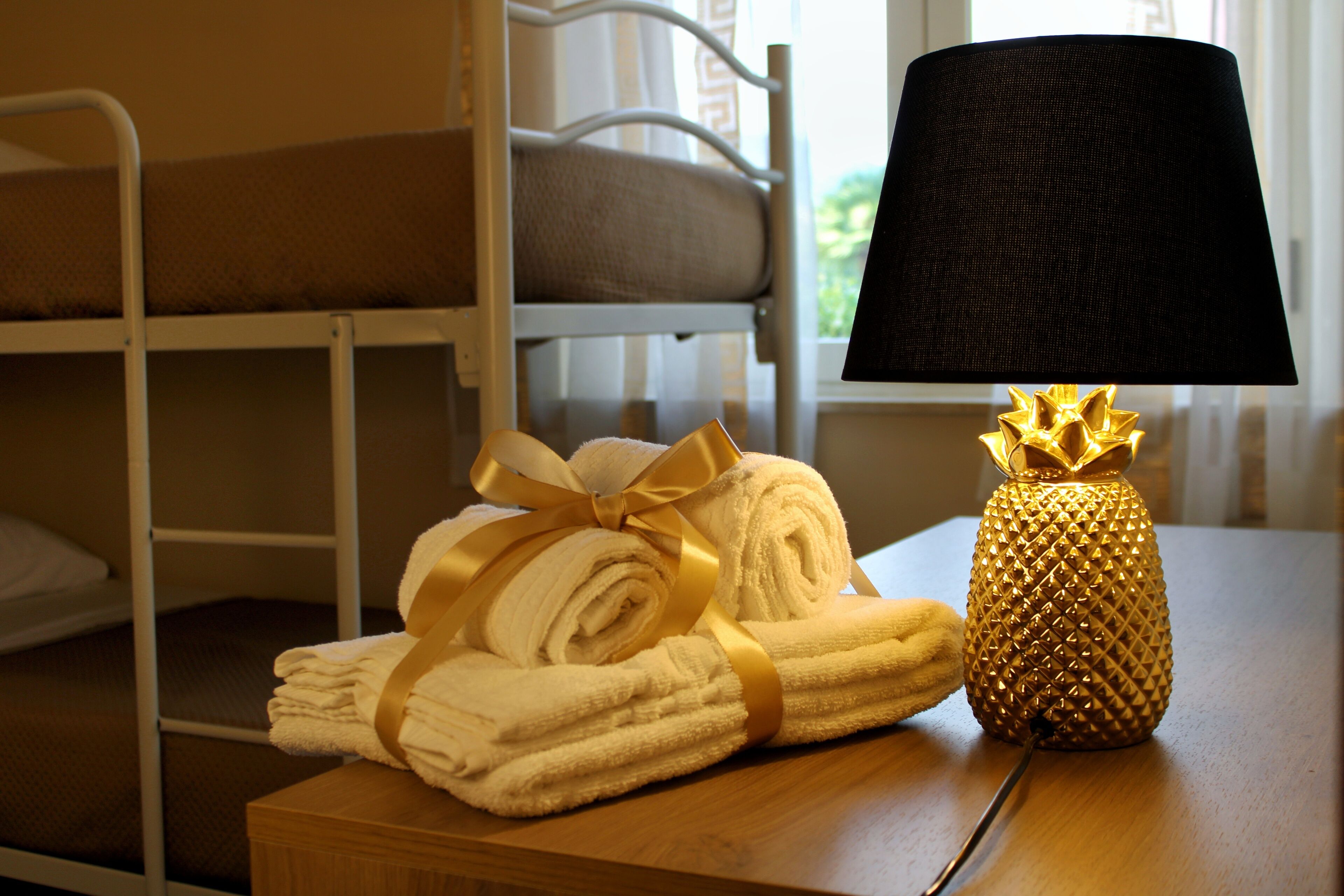 A neatly made bed with a white headboard, a rolled-up towel with a gold ribbon, and a lamp with a textured lampshade on a wooden table.