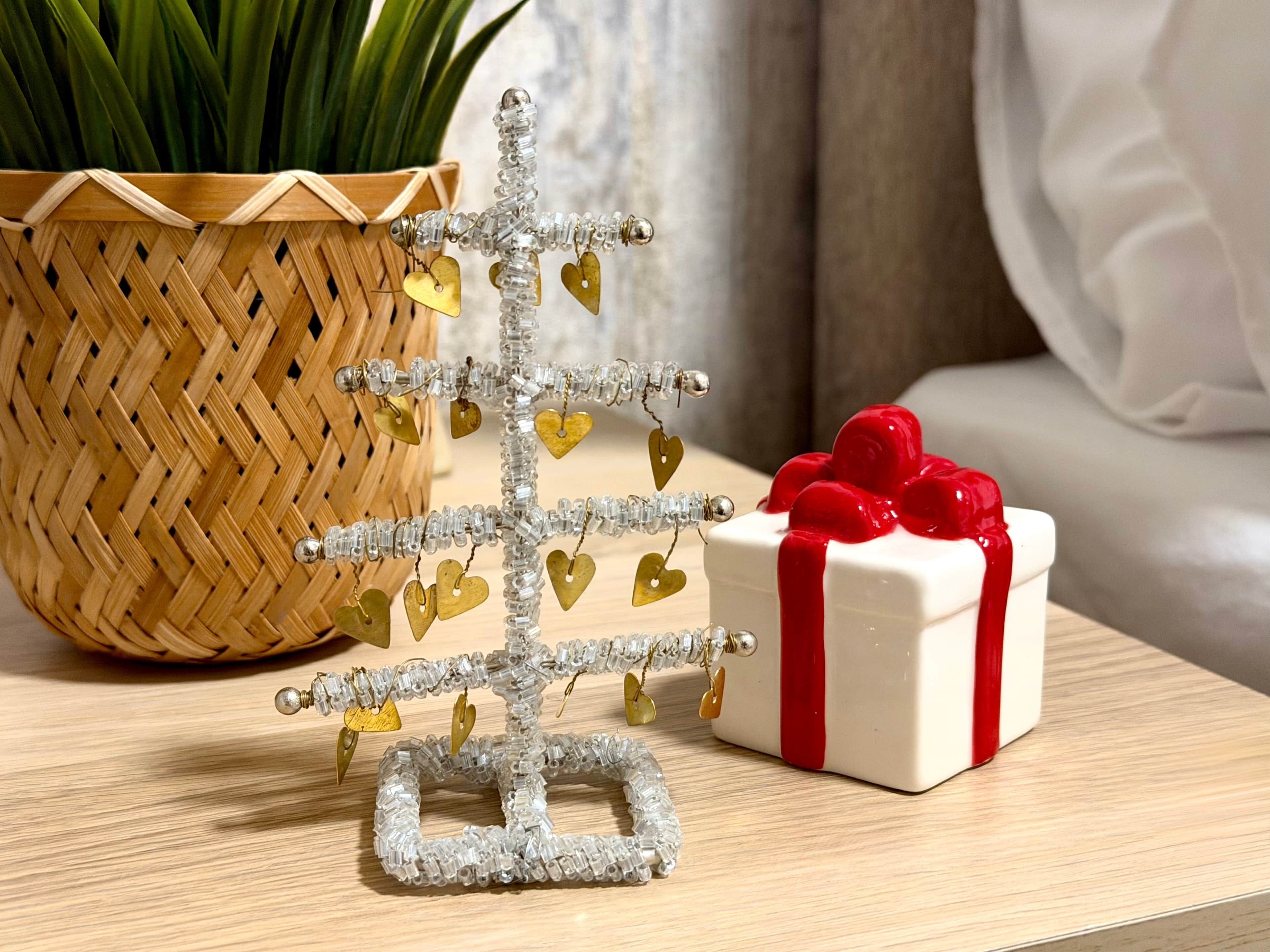 Un petit arbre décoratif en argent, orné de petits cœurs, une boîte cadeau blanche avec un ruban rouge et un panier tressé contenant des plantes vertes.