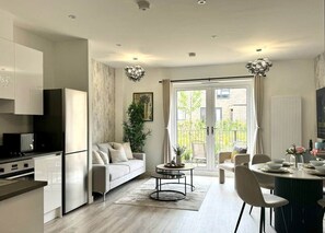 Eine moderne Küche mit weißen Schränken, ein Essbereich mit rundem Tisch und ein Wohnzimmer mit Sofa und Pflanze.