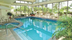 Una piscina interior con paredes de vidrio transparente, rodeada de sillas blancas y plantas en macetas.