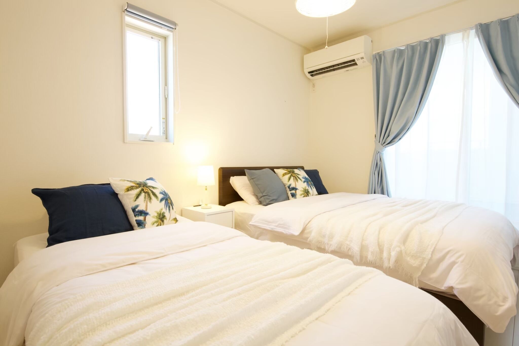 A hotel room with two beds, a window, an air conditioning unit, and a nightstand with a lamp.