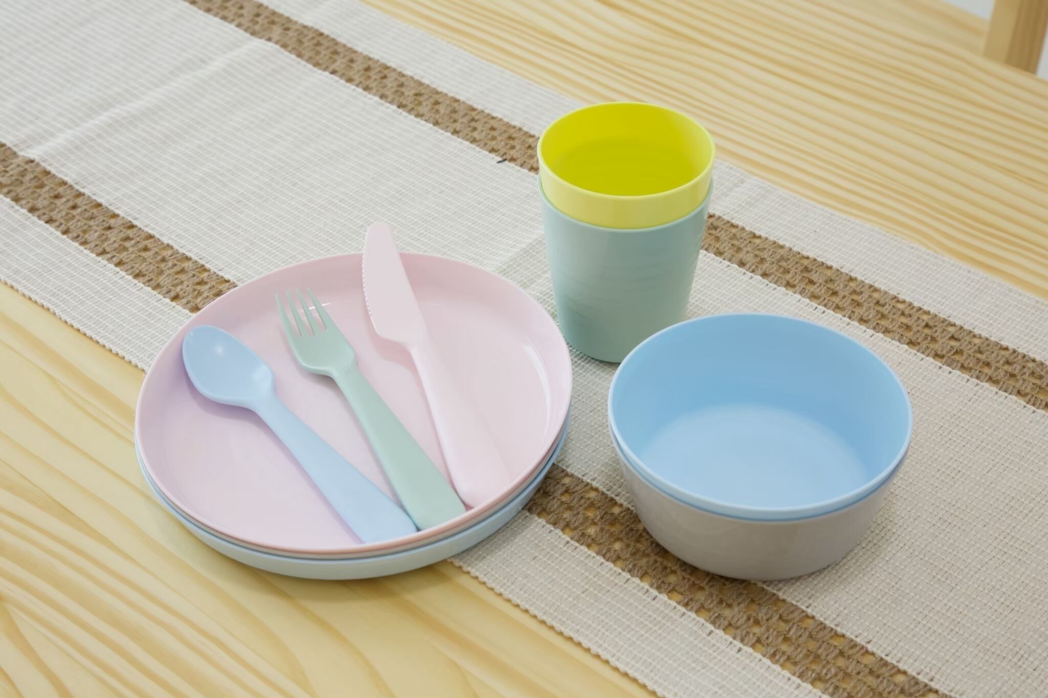 A table with a striped tablecloth, a pink plate with a fork and knife, a yellow cup, and a blue bowl.