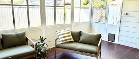 A covered porch with wooden flooring, two green cushioned sofas, a small round table with two wine glasses and a bottle, and a potted plant.