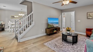 A spacious living room with a wooden staircase, a ceiling fan, a flat-screen TV, and a dining area with a white table and chairs.