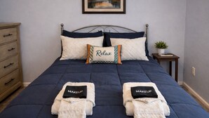 Un lit bien fait, recouvert d’une couvre-lit bleu foncé, avec deux oreillers dont l’un porte le mot « Relax » et deux serviettes sur lesquelles est écrit « MAQUILLAGE ».