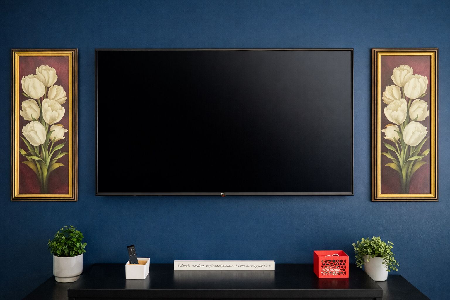 Habitación con un televisor de pantalla plana grande montado en una pared azul, flanqueado por dos cuadros con flores blancas. Hay una mesa negra con una planta en maceta, un control remoto y un cartel con un mensaje humorístico.