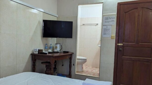 A hotel room with a bed, a wooden bedside table with a TV, a mirror, a bathroom with a toilet, and a wooden door. - Empire House Phnom Penh (Phnom Penh)