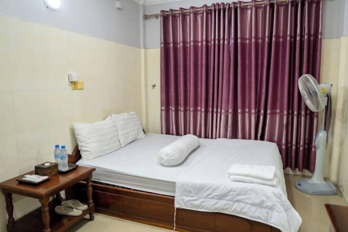 A hotel room with a bed, a bedside table with a fan, a water bottle, and slippers.