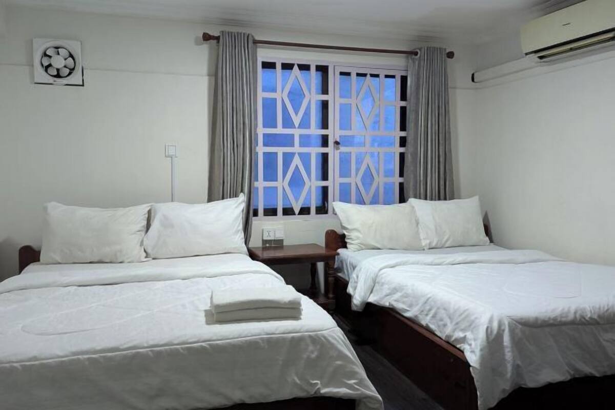 A hotel room with two beds, a window with curtains, an air conditioning unit, and a wall-mounted fan.