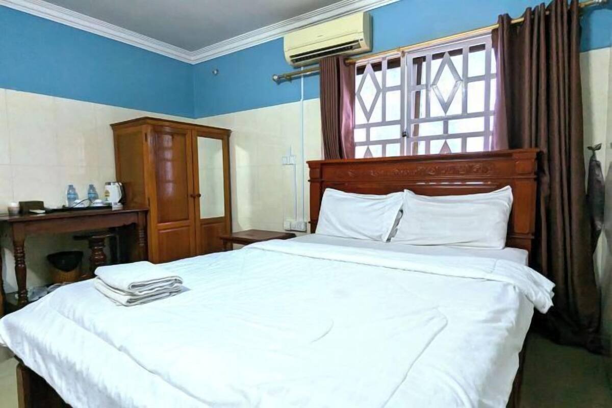 A hotel room with a bed, a wooden wardrobe, a desk with a chair, and a window with curtains.