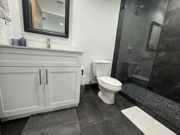 A bathroom with a white vanity, a toilet, a shower with dark tiles, and a mirror above the vanity.