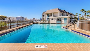 A swimming pool with a 7 ft 6 in depth sign, surrounded by a fence, and adjacent to a building with balconies. - Charming Walkable 1BR condo with Canal View and Heated Pool (Fort Myers Beach)