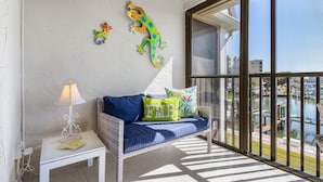 A balcony with a wicker sofa, a small table with a lamp, decorative wall art, and a view of a marina. - Charming Walkable 1BR condo with Canal View and Heated Pool (Fort Myers Beach)