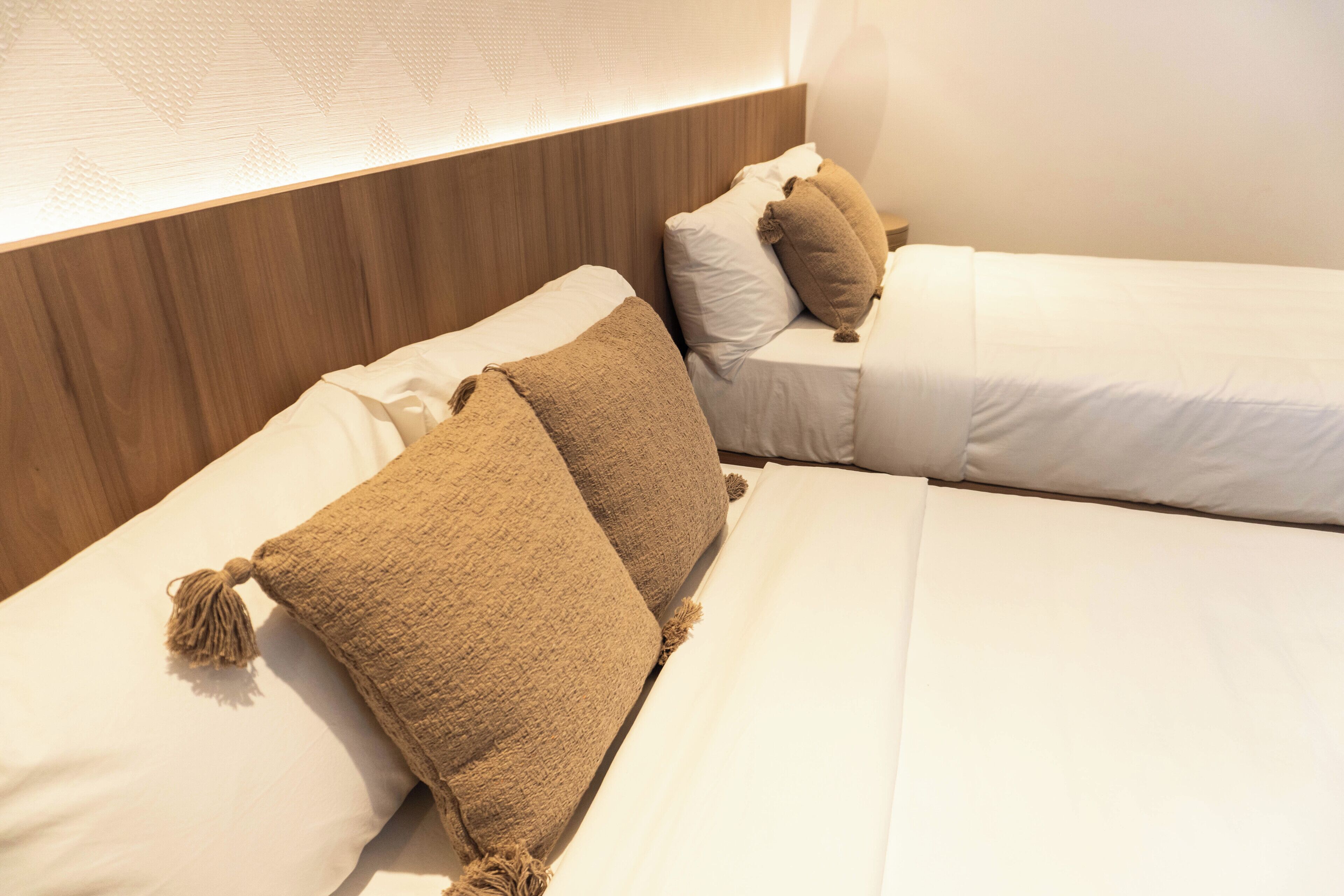 A hotel room with a wooden headboard, two beds with white bedding, and beige throw pillows.
