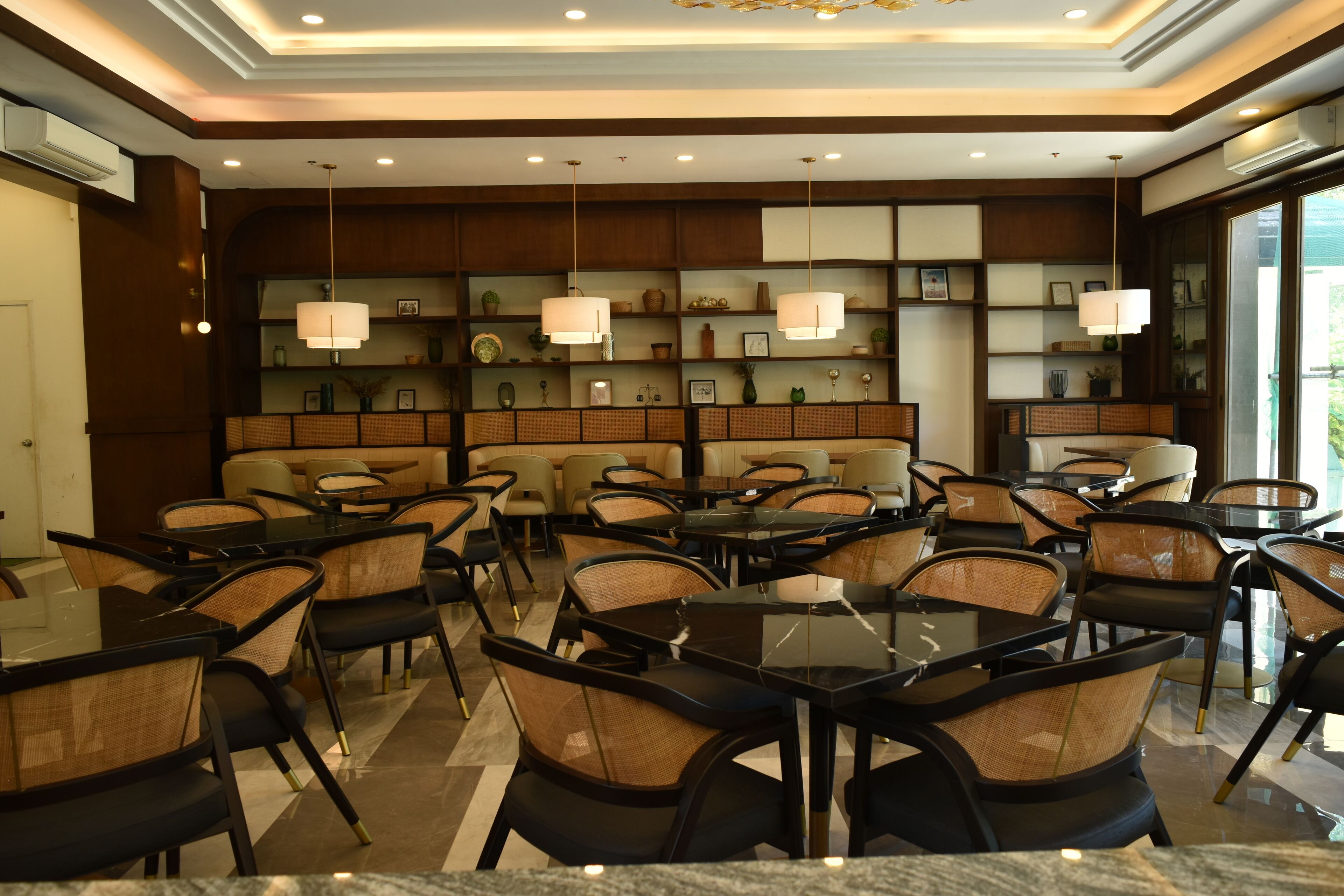A hotel dining area with tables and chairs, a decorative wall with shelves, and pendant lighting.