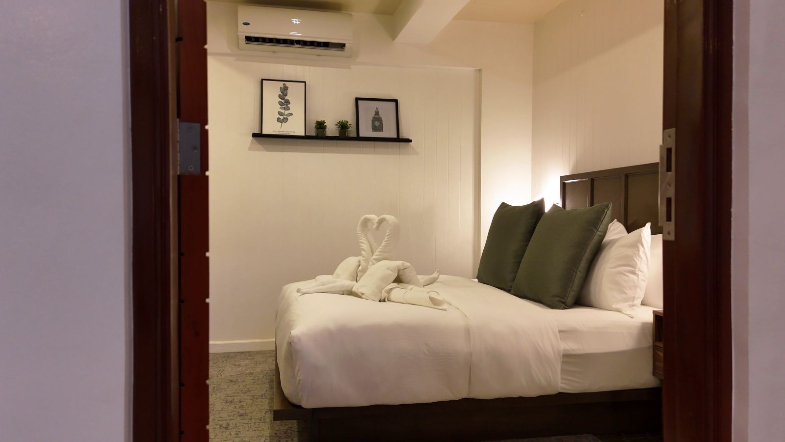 A hotel room with a neatly made bed, a headboard, and a shelf with framed pictures.