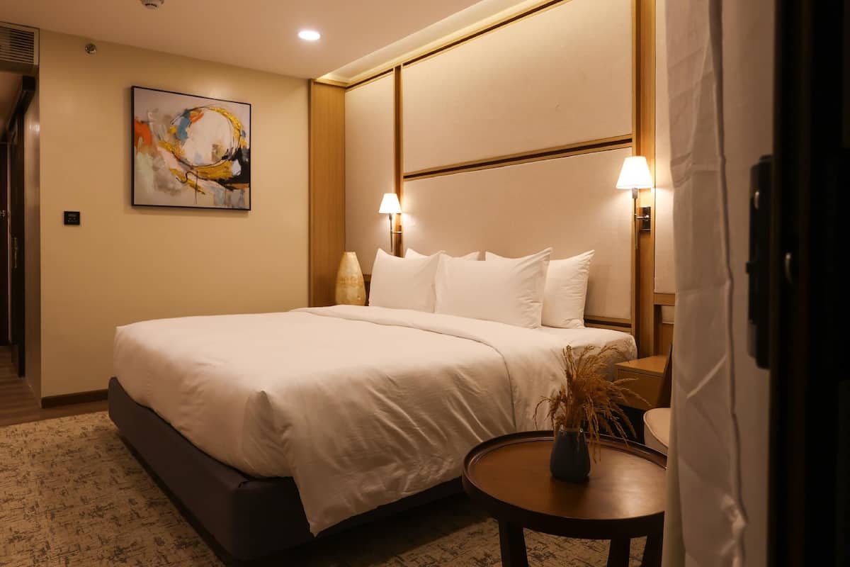 Une chambre d’hôtel avec un grand lit, deux lampes de chevet, un tableau au mur et une petite table sur laquelle est posé un vase.