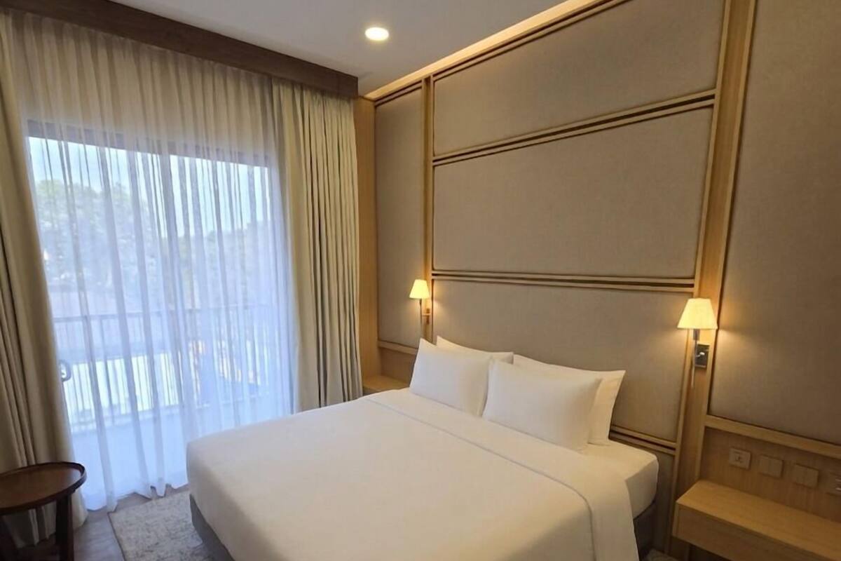 Une chambre d’hôtel avec un grand lit, une table de chevet, une lampe et une vue sur l’extérieur à travers des rideaux légers.