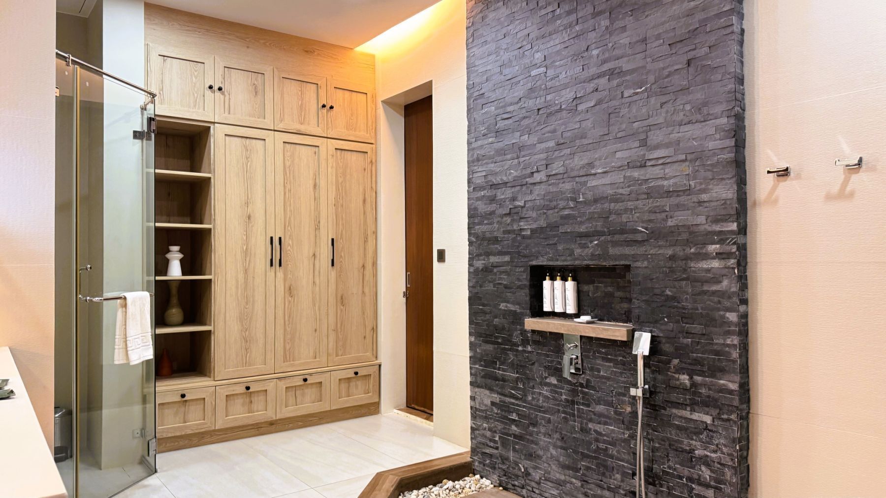 Un baño con un armario de madera, una cabina de ducha de vidrio, una pared de piedra y un estante empotrado con artículos de aseo.