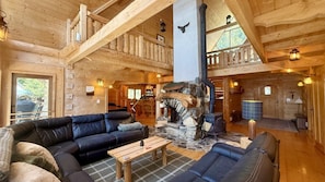 A log cabin interior with a stone fireplace, leather sofas, and a wooden staircase.
