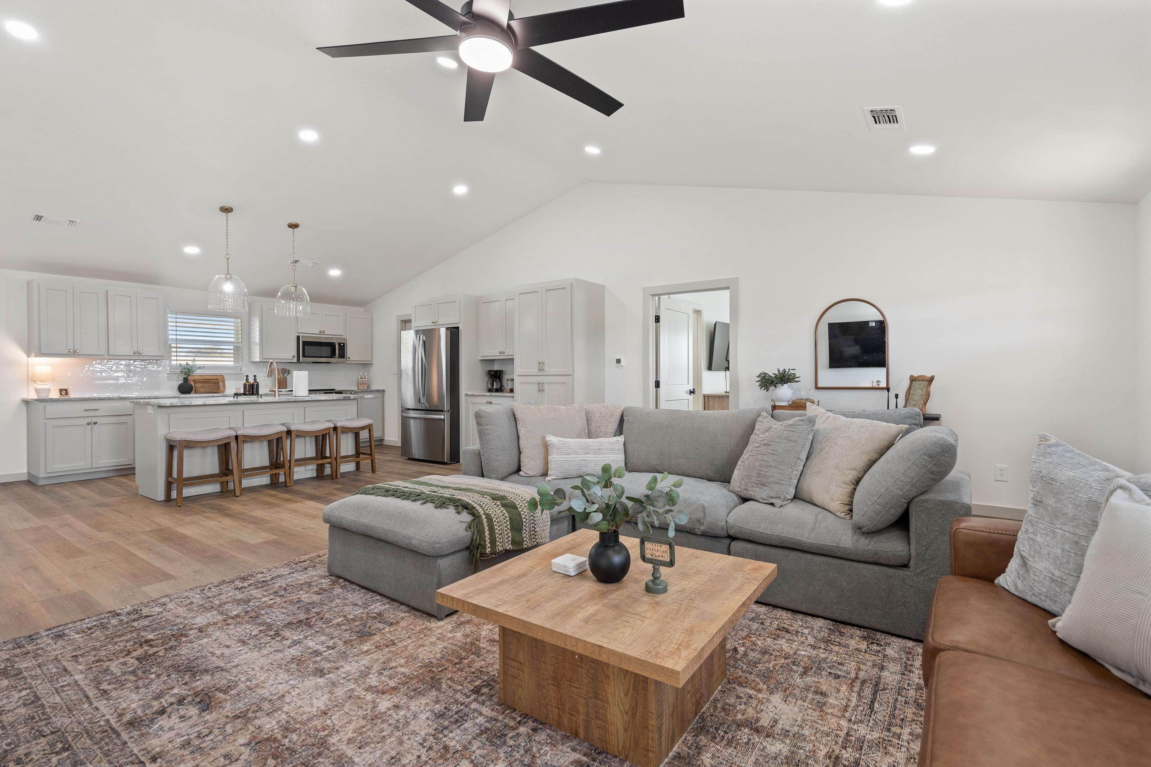 Eine moderne Küche mit weißen Schränken, einer zentralen Kochinsel mit Sitzgelegenheiten, einem Essbereich mit grauem Sofa und Couchtisch sowie einem Deckenventilator.