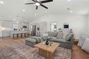 Eine moderne Küche mit weißen Schränken, einer zentralen Kochinsel mit Sitzgelegenheiten, einem Essbereich mit grauem Sofa und Couchtisch sowie einem Deckenventilator.