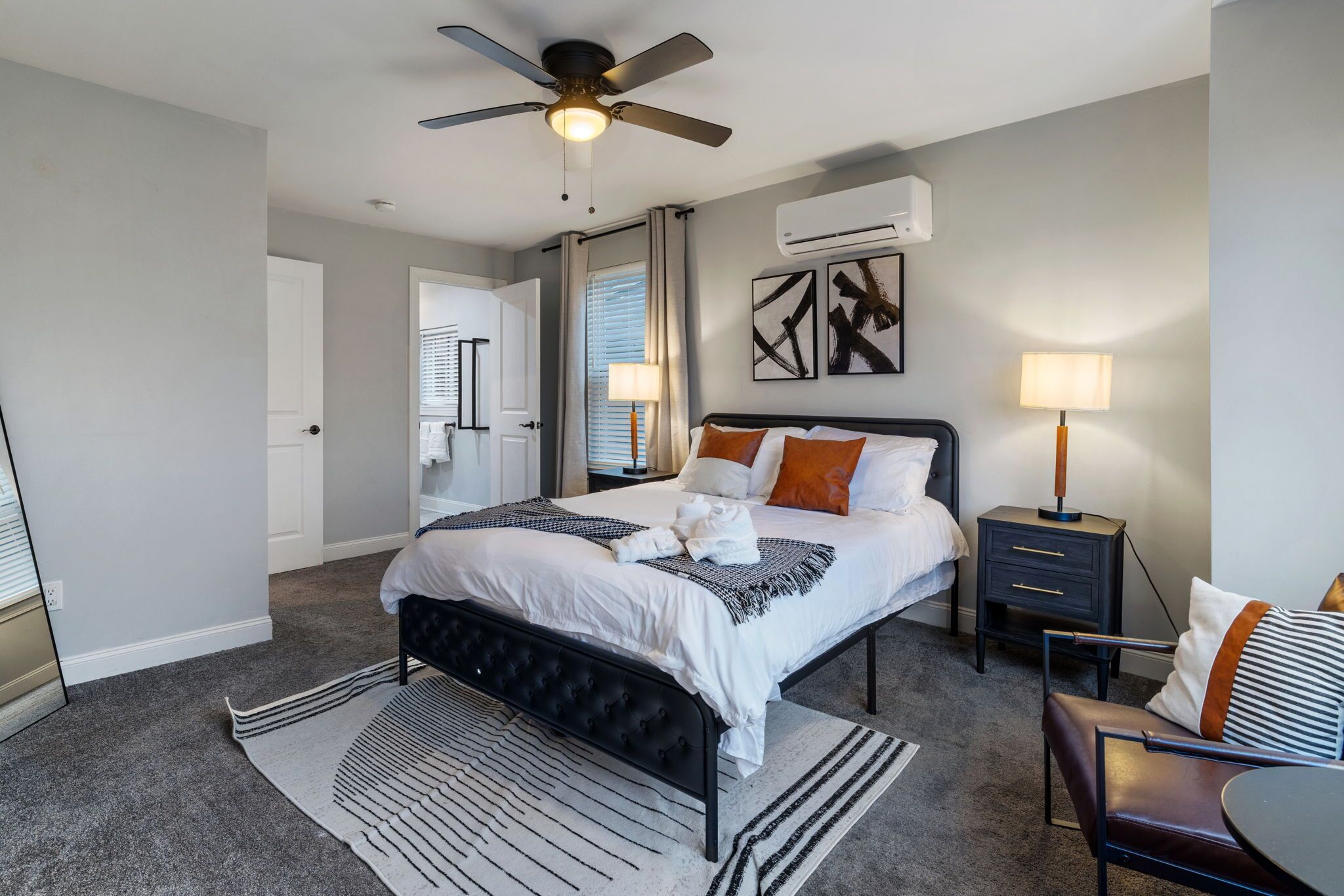 A bedroom with a bed, bedside tables, a ceiling fan, an air conditioning unit, and a view of the adjacent room.