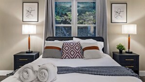 A neatly made bed with a patterned throw, two bedside lamps, a window with a view of greenery, and framed artwork on the wall. - Modern 3BR Skyline Retreat w/ Rooftop Deck + Free Parking + Views (Pittsburgh)