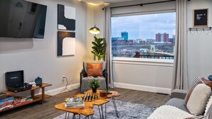 A modern living room with a large window, a sofa, a coffee table, a TV, and a bookshelf. - Modern 3BR Skyline Retreat w/ Rooftop Deck + Free Parking + Views (Pittsburgh)
