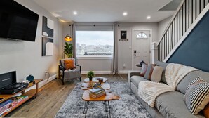 A modern living room with a sofa, a coffee table, a TV, and a staircase. - Modern 3BR Skyline Retreat w/ Rooftop Deck + Free Parking + Views (Pittsburgh)