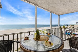 Een ronde tafel met een geweven placemat, omringd door rieten stoelen, met uitzicht op een strand en een helder zicht op de oceaan en verre gebouwen.
