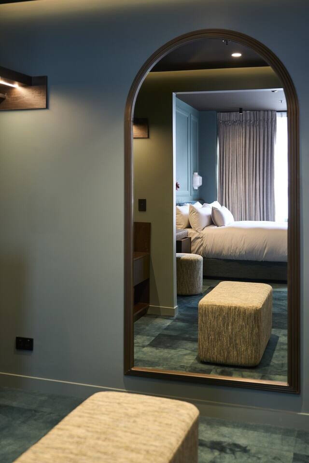 Hotelkamer met een grote spiegel die een bed, nachtkastje en raam met gordijnen weerspiegelt.
