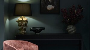 Una stanza con una poltrona rosa, un tavolino consolle scuro, una lampada, un quadro incorniciato e un vaso con fiori.