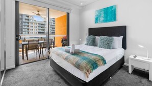A modern hotel room with a large bed, a ceiling fan, a view of the city through a sliding door, and a small table with chairs.