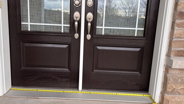 A dark wooden double door with a window, a doorbell, and a measuring tape on the ground.