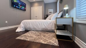 A bedroom with a bed, a TV mounted on the wall, a bedside table with a lamp, and a window with blinds.