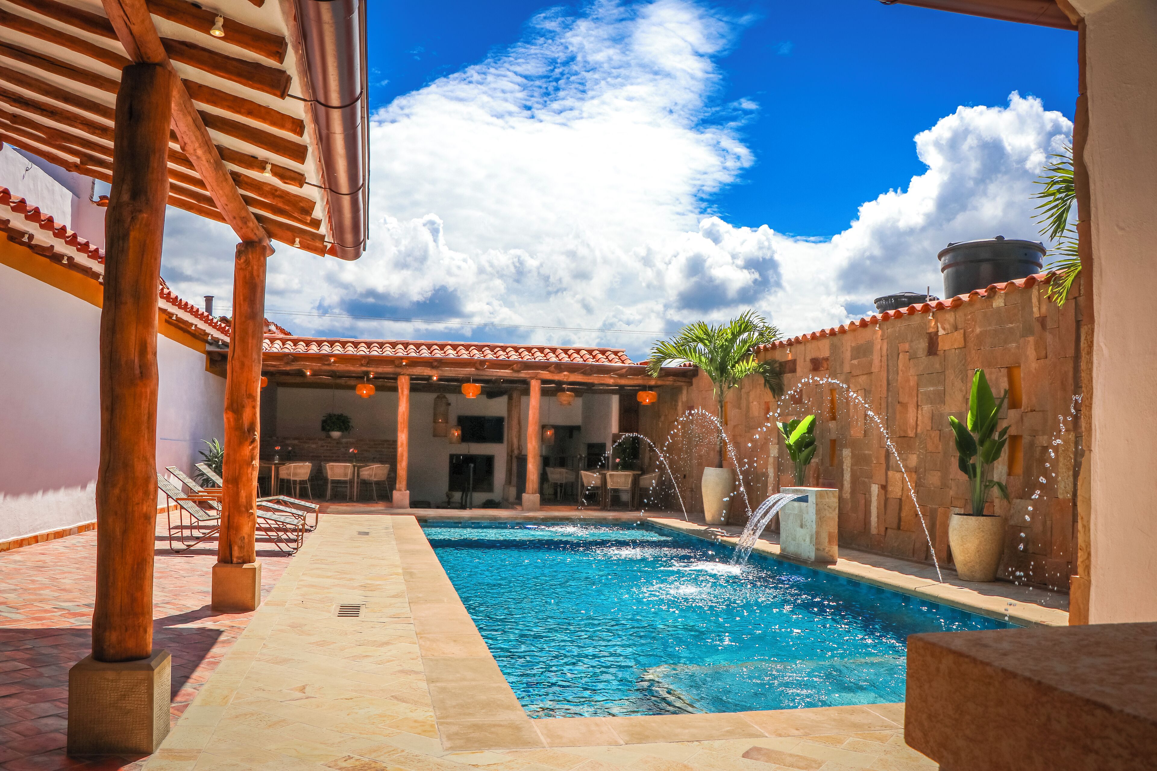 A swimming pool with water jets, surrounded by a covered patio with wooden pillars and a tiled floor.