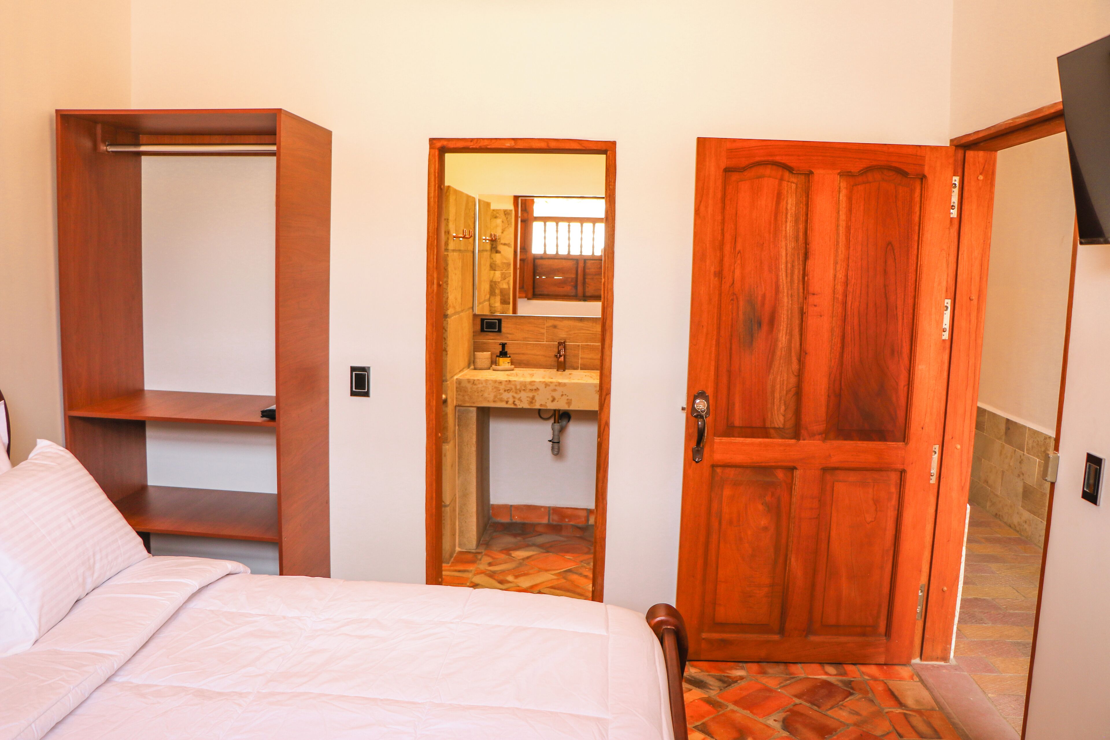 Een slaapkamer met een bed, een houten garderobe, een badkamer met een wastafel en spiegel, en een houten deur.