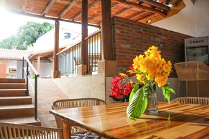 Una mesa de madera con un jarrón de flores, sillas de mimbre y un muro de ladrillo al fondo.