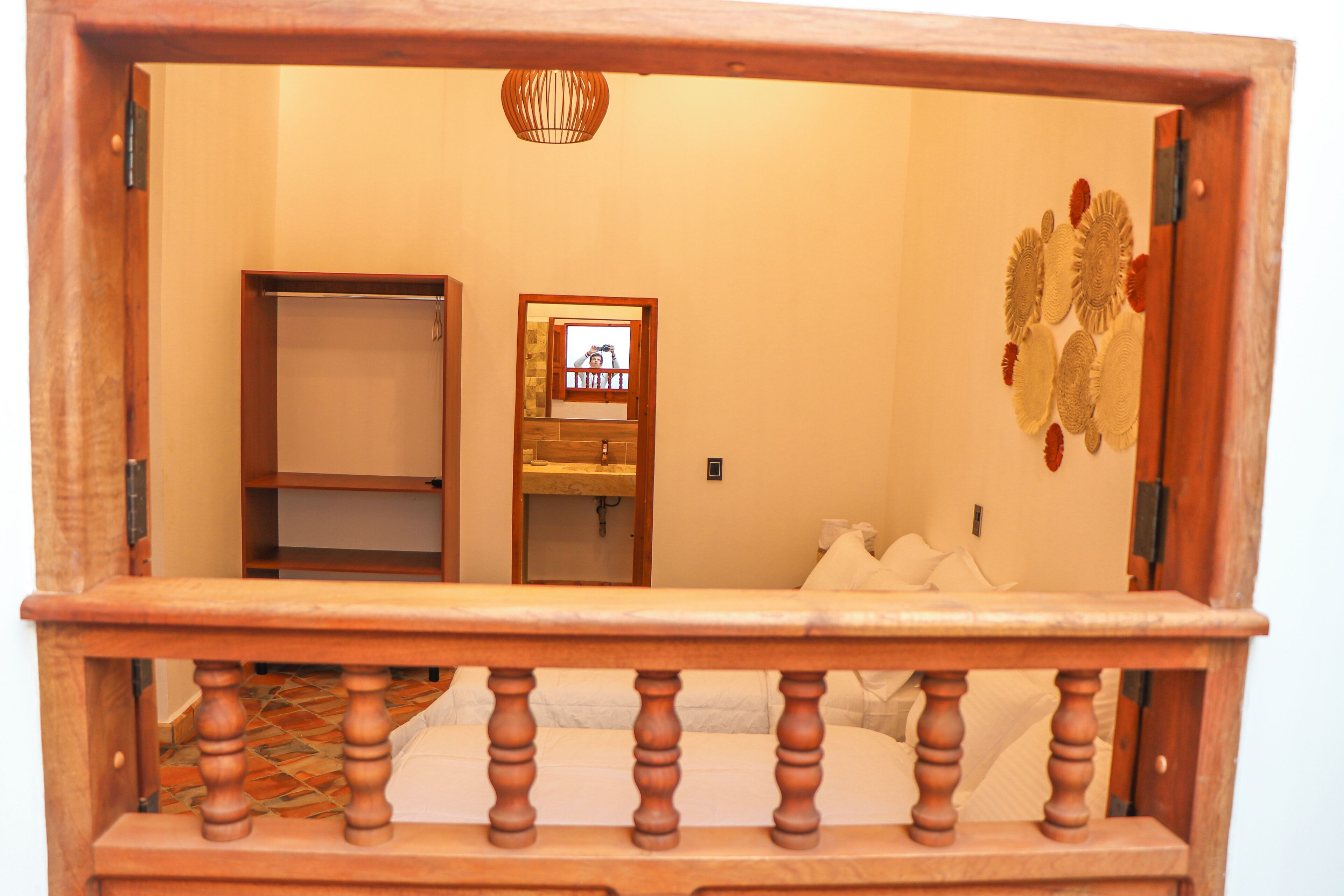 A bedroom with a wooden headboard, a bed with white bedding, a wooden wardrobe, and a mirror reflecting a bathroom with a sink and a towel rack.
