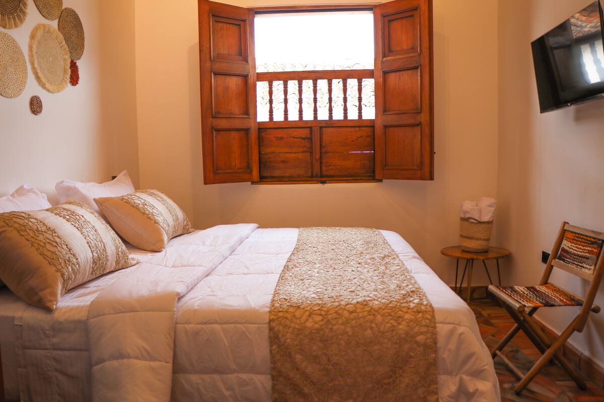 A bedroom with a bed, a window with wooden shutters, a television on the wall, and a small table with a chair.