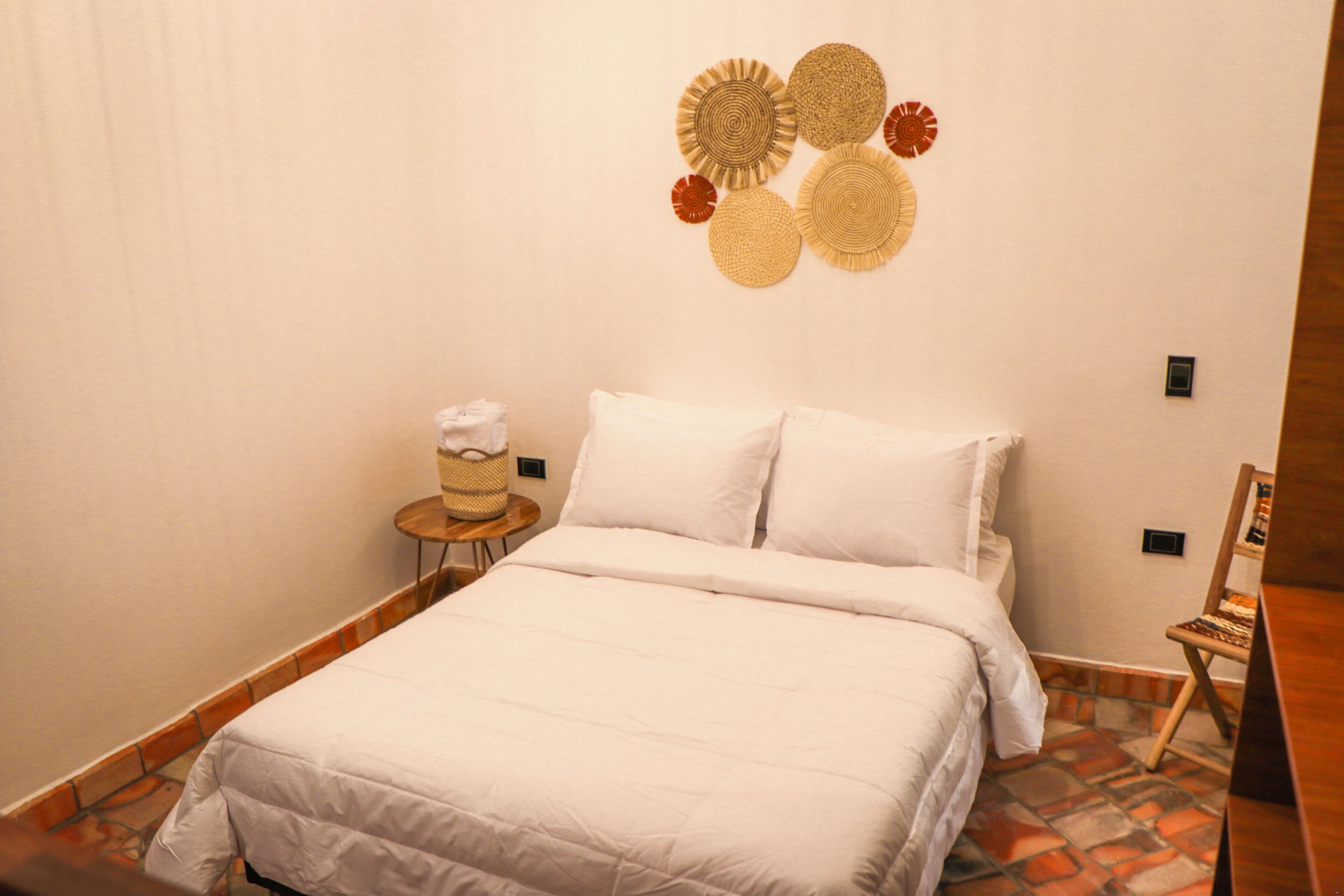 A neatly made bed with white bedding, a small round table with a basket, and a wall-mounted decorative item.